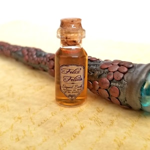 May include: A miniature glass bottle with a cork stopper, filled with a golden liquid, labeled "Felix Felicis". Beside it is a wand with a brown and copper-colored handle and a teal-colored crystal at the end. The items rest on a parchment-like surface.