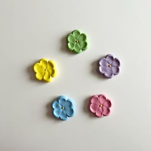 May include: Five colourful flower-shaped magnets in yellow, green, purple, blue, and pink. Each magnet has a small gold centre.