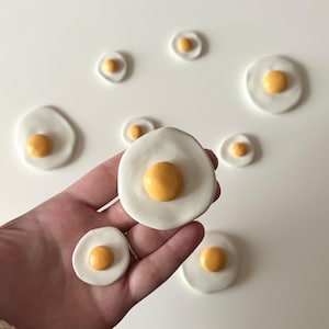 May include: A hand holding several ceramic fried eggs with white 'whites' and yellow 'yolks'. Several other ceramic eggs are scattered on a white surface. The eggs vary in size and are handmade.