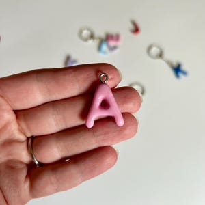 Op de afbeelding: Een lichtroze letter A-bedel met een gladde, glanzende afwerking en een zilverkleurige metalen ring aan de bovenkant. De bedel heeft een afgeronde, bolle vorm en wordt getoond naast verschillende andere kleine, onscherpe letterbedels.