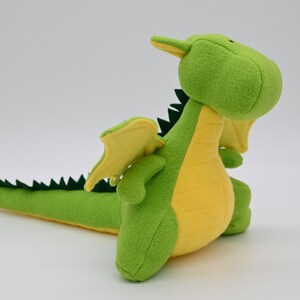 May include: A green and yellow plush dragon toy with a long tail and wings. The dragon is sitting up with its head tilted back.