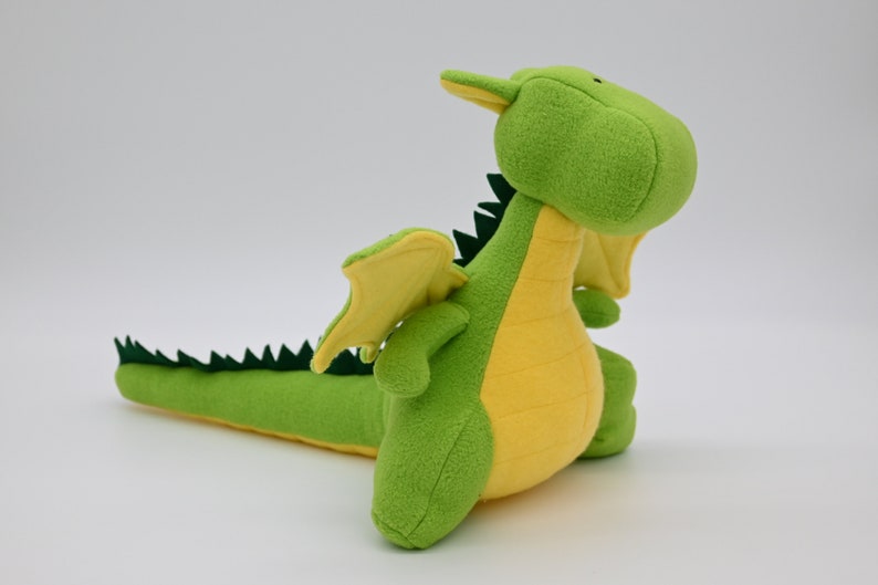 May include: A green and yellow plush dragon toy with a long tail and wings. The dragon is sitting up with its head tilted back.
