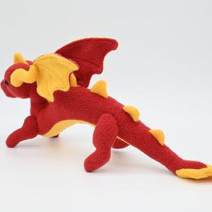 May include: A plush red dragon toy with yellow accents. The dragon has yellow wings, horns, and spikes along its back. The toy is made of a soft material and is posed in a running position. The dragon is a fantasy-themed item.