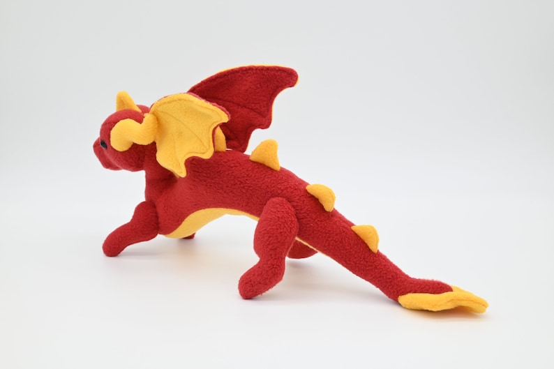 May include: A plush red dragon toy with yellow accents. The dragon has yellow wings, horns, and spikes along its back. The toy is made of a soft material and is posed in a running position. The dragon is a fantasy-themed item.