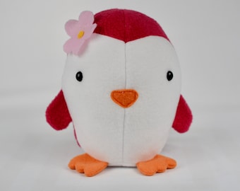 May include: A plush penguin toy with a white body, red head and wings, and orange feet and beak. A pink flower with a yellow center adorns the penguin's head. The penguin has black button eyes and is made of soft fabric.