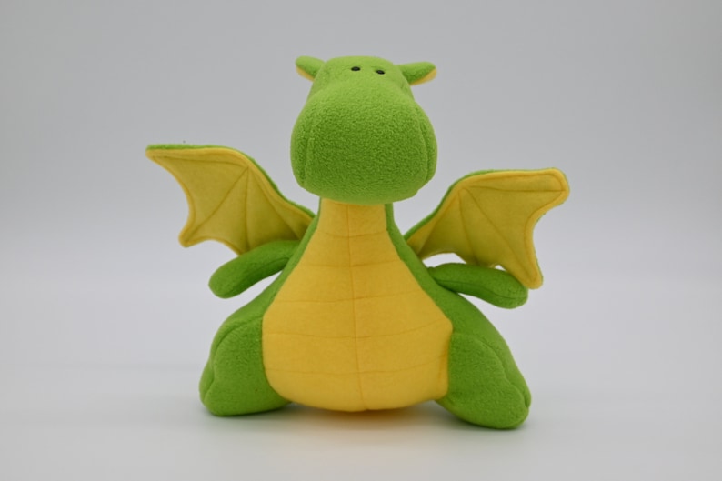 May include: A green and yellow plush dragon toy with wings. The dragon has a friendly face and is sitting on a white surface.