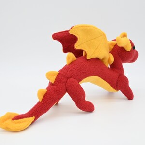 May include: A plush dragon toy in red and yellow. The dragon has a long tail, wings, and horns. The body is red, with yellow accents on the wings, horns, and tail. The dragon is posed on a white background.