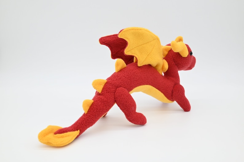 May include: A plush dragon toy in red and yellow. The dragon has a long tail, wings, and horns. The body is red, with yellow accents on the wings, horns, and tail. The dragon is posed on a white background.