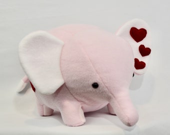 May include: A pink plush elephant with white ears and a red heart design on one ear. The elephant has black eyes and a small, curled trunk.