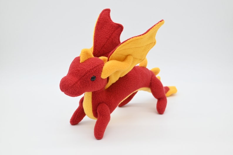 May include: A plush dragon toy in red and yellow. The dragon has large, yellow wings, a long tail, and small black eyes. The toy is made of soft fabric and is designed to look like a friendly dragon.