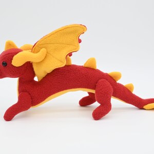 May include: A plush red dragon toy with yellow wings, horns, and accents. The stuffed animal has a long tail and is posed in a running position. The dragon is made of soft fabric and has black button eyes.