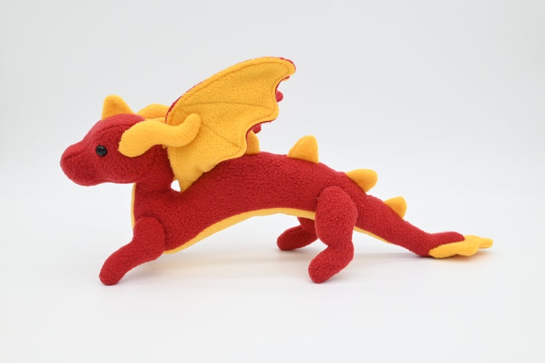 May include: A plush red dragon toy with yellow wings, horns, and accents. The stuffed animal has a long tail and is posed in a running position. The dragon is made of soft fabric and has black button eyes.