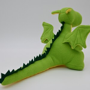 May include: Green plush dragon toy with yellow accents. The dragon has wings, a long tail with green spikes, and a friendly face.