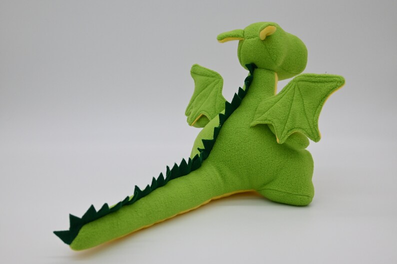 May include: Green plush dragon toy with yellow accents. The dragon has wings, a long tail with green spikes, and a friendly face.