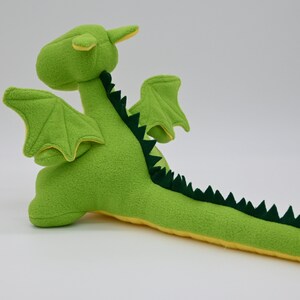 May include: Green plush dragon toy with yellow accents. The dragon has wings, a long tail with green spikes, and a small horn on its head.