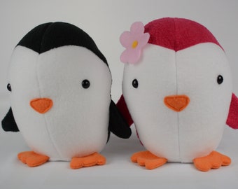 May include: Two plush penguin toys, one black and white and one pink and white, with orange beaks and feet. The pink penguin has a pink flower on its head.