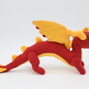 May include: A plush red dragon toy with yellow wings, horns, and tail. The stuffed animal has a detailed design, with a long body and four legs. The dragon is posed on a white background.