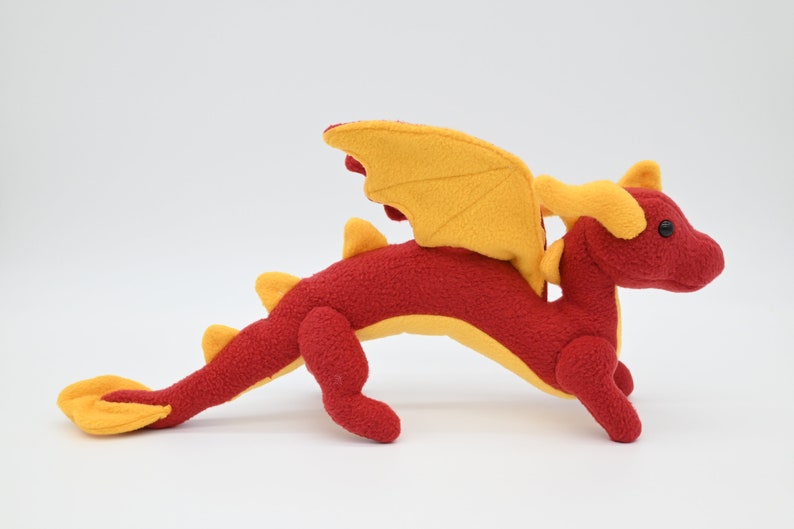 May include: A plush red dragon toy with yellow wings, horns, and tail. The stuffed animal has a detailed design, with a long body and four legs. The dragon is posed on a white background.