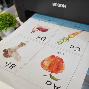 May include: A set of four printable flashcards featuring watercolour illustrations of an apple, a bird, a deer, and a carrot. Each card has a letter and the word for the image, such as "a apple" and "b bird".