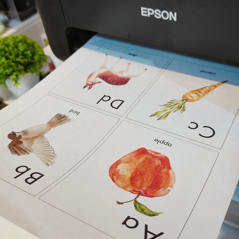 May include: A set of four printable flashcards featuring watercolour illustrations of an apple, a bird, a deer, and a carrot. Each card has a letter and the word for the image, such as "a apple" and "b bird".