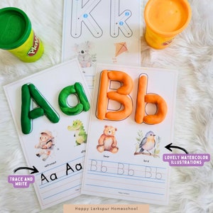 May include: A printable worksheet for learning the letters A and B. The worksheet features a green playdough letter A, an orange playdough letter B, and illustrations of an astronaut, alligator, bear, and bird. The text "TRACE AND WRITE" is in a purple box at the bottom left corner of the worksheet. The text "LOVELY WATERCOLOR ILLUSTRATIONS" is in a purple box at the bottom right corner of the worksheet. The text "Happy Larkspur Homeschool" is at the bottom center of the worksheet.