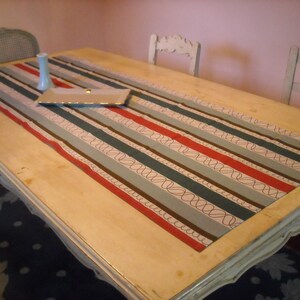 May include: A white wooden table with a striped table runner in shades of blue, gray, brown, and red. The table runner has a pattern of swirls and lines. The table is set with a white rectangular tray.