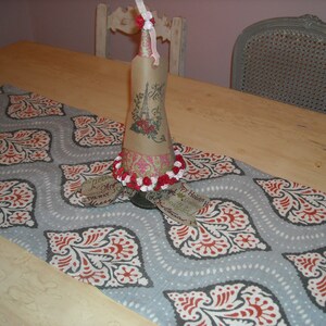 May include: A gray and red patterned table runner with a decorative bottle wrapped in paper and decorated with red and white flowers. The bottle has a tag that says "My Holiday Home".