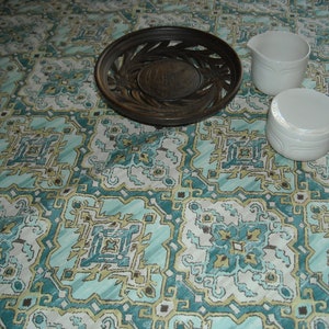 May include: Two white ceramic creamer and sugar bowls with a decorative design on a teal and brown patterned tablecloth. A wooden decorative tray with a woven design is also on the tablecloth.