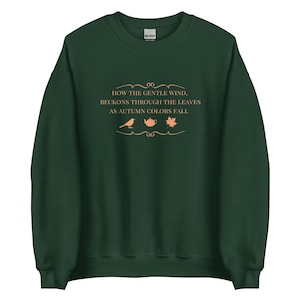 Op de afbeelding: Een donkergroene sweatshirt met de tekst "HOW THE GENTLE WIND, BECKONS THROUGH THE LEAVES AS AUTUMN COLORS FALL" in lichtoranje. Het ontwerp bevat een vogel, een theepot en een blad.