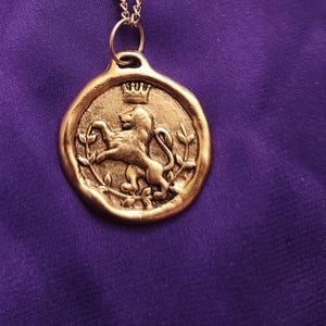May include: A gold-toned necklace featuring a circular pendant with an embossed lion figure wearing a crown, flanked by two leafy branches. The pendant hangs from a delicate gold-toned chain with small, evenly spaced decorative metal beads.