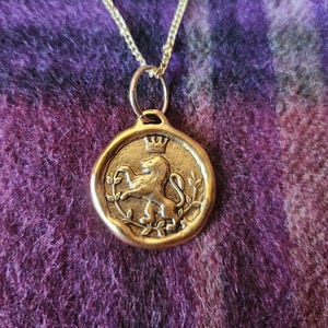 May include: A gold-toned circular pendant necklace featuring a raised relief of a lion standing on its hind legs beneath a small crown, framed by thin leafy branches, suspended from a delicate gold-toned metal chain link necklace.