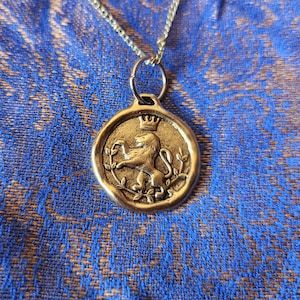 May include: A gold-toned metal pendant necklace featuring a circular wax seal design. The pendant displays a raised relief of a lion standing on its hind legs, topped with a small crown, and surrounded by thin leafy branches on a textured surface.