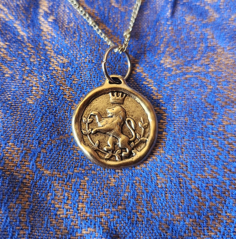 May include: A gold-toned metal pendant necklace featuring a circular wax seal design. The pendant displays a raised relief of a lion standing on its hind legs, topped with a small crown, and surrounded by thin leafy branches on a textured surface.