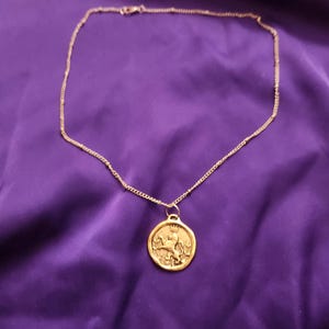 May include: A gold-toned necklace featuring a thin chain and a circular pendant. The pendant displays a raised relief design of a lion wearing a crown, standing on its hind legs, surrounded by a thin border. The metal has a matte, textured finish.