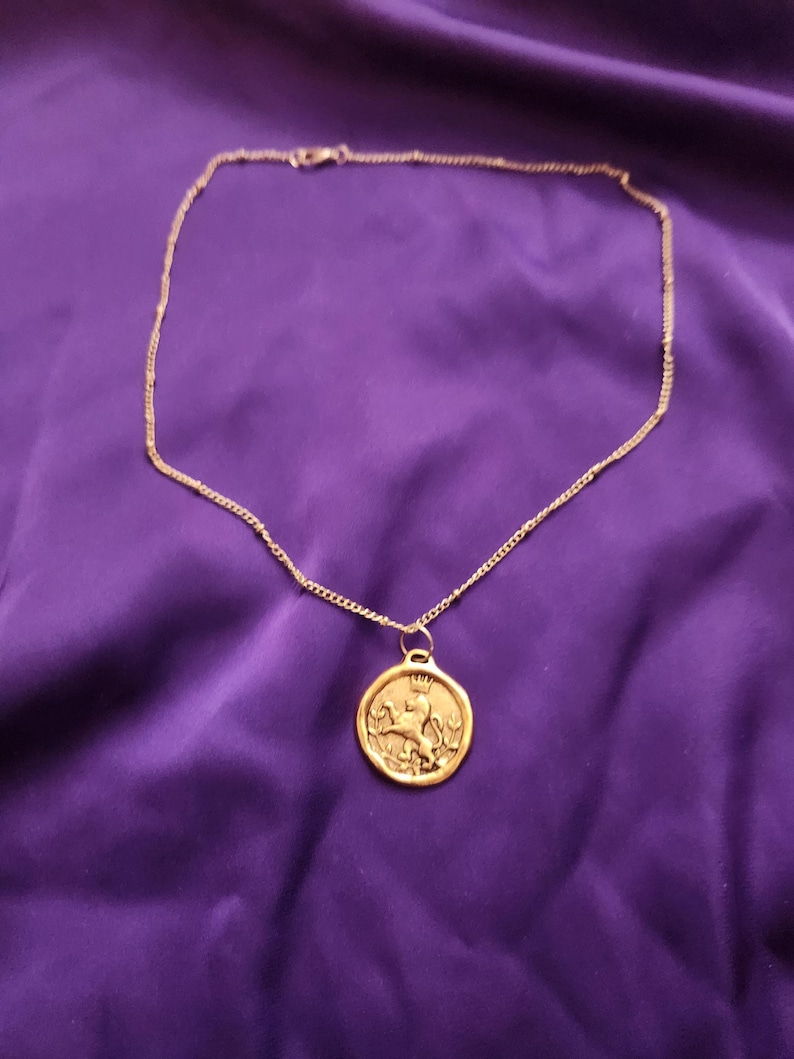 May include: A gold-toned necklace featuring a thin chain and a circular pendant. The pendant displays a raised relief design of a lion wearing a crown, standing on its hind legs, surrounded by a thin border. The metal has a matte, textured finish.