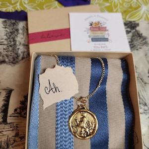 May include: A gold-toned circular pendant featuring a lion wearing a crown, suspended from a delicate gold chain. The necklace rests on a blue and beige striped fabric inside a square cardboard box, accompanied by a small tag with the text Ah.