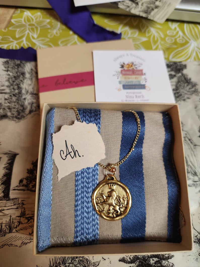 May include: A gold-toned circular pendant featuring a lion wearing a crown, suspended from a delicate gold chain. The necklace rests on a blue and beige striped fabric inside a square cardboard box, accompanied by a small tag with the text Ah.
