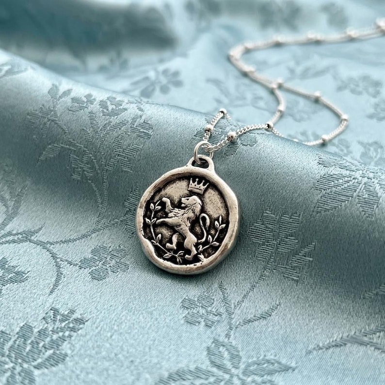 May include: A silver-toned circular pendant necklace featuring a raised relief of a lion wearing a crown, surrounded by leafy branches. The pendant hangs from a delicate silver-toned chain with small, evenly spaced spherical beads.