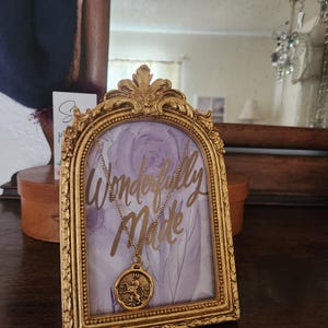 May include: A gold-toned necklace with a round pendant featuring a lion emblem, displayed on a decorative gold arched frame. The frame contains a light purple floral background with the gold script text Wonderfully Made.
