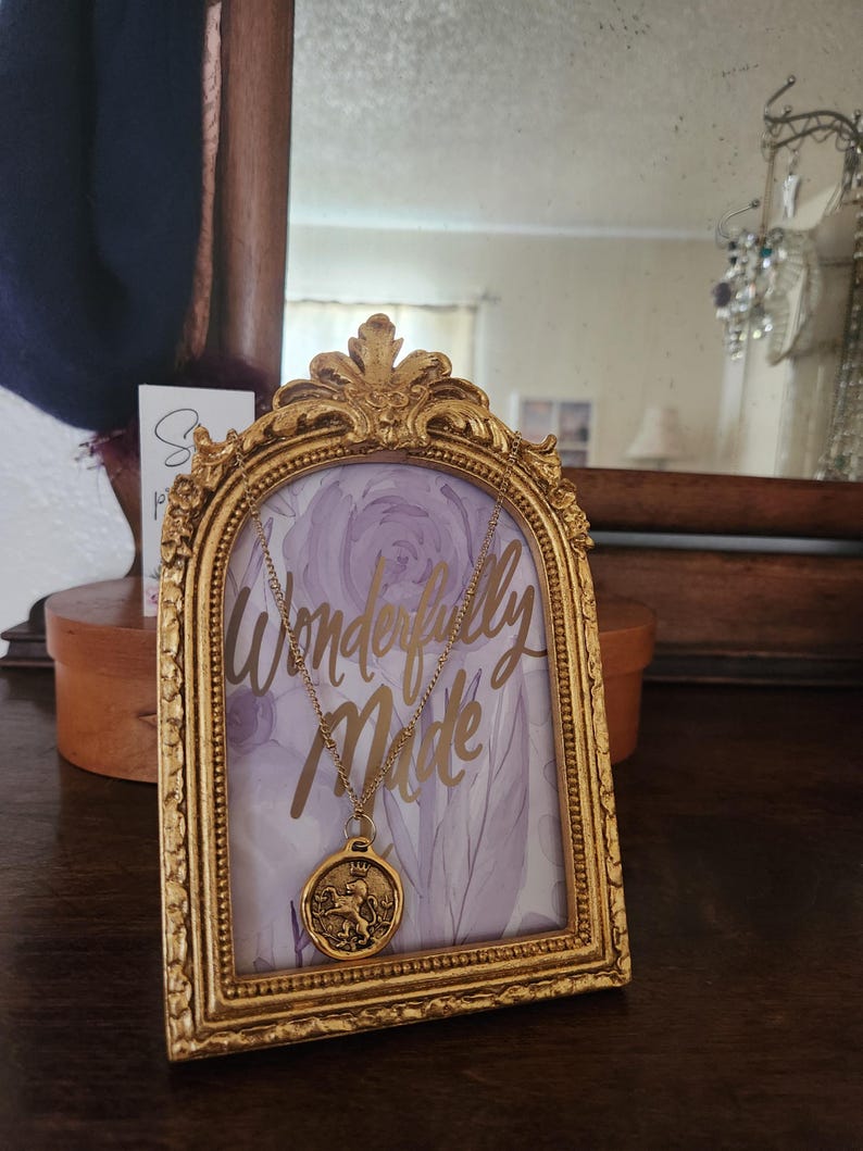 May include: A gold-toned necklace with a round pendant featuring a lion emblem, displayed on a decorative gold arched frame. The frame contains a light purple floral background with the gold script text Wonderfully Made.