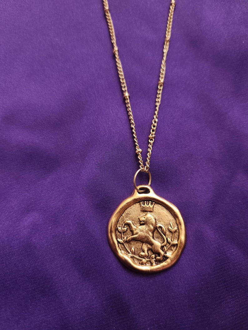 May include: A gold-toned necklace featuring a circular pendant with an embossed lion figure wearing a crown, flanked by two leafy branches. The pendant hangs from a delicate gold-toned chain with small, evenly spaced decorative metal beads.