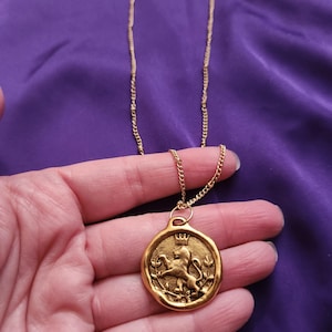 May include: A gold-toned necklace featuring a circular pendant with an embossed design of a lion wearing a crown, surrounded by leafy branches. The pendant has an irregular, wax-seal style edge and hangs from a delicate gold-toned metal chain.