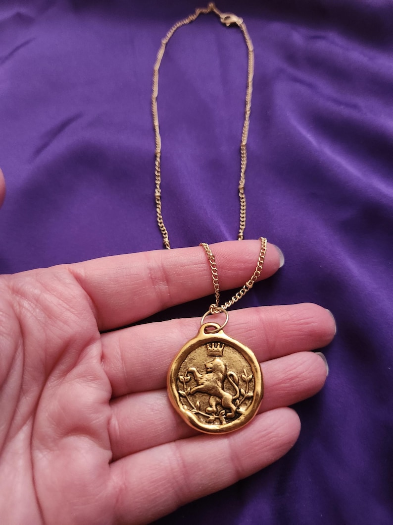 May include: A gold-toned necklace featuring a circular pendant with an embossed design of a lion wearing a crown, surrounded by leafy branches. The pendant has an irregular, wax-seal style edge and hangs from a delicate gold-toned metal chain.