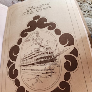 May include: A tan paper souvenir menu featuring a brown ink illustration of the Steamboat Delta Queen inside an ornate oval frame. The text Steamboat Delta Queen is printed at the top, and the words Souvenir Menu are printed at the bottom.