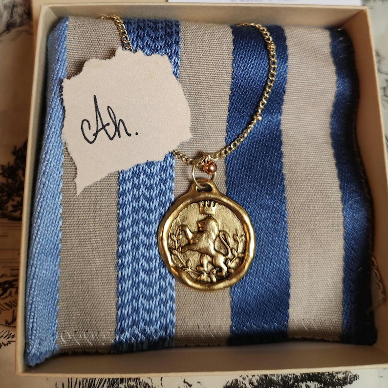 May include: A gold-toned circular pendant featuring a raised lion design with a crown, suspended from a thin gold-toned chain. The pendant rests on a blue and beige striped fabric inside a box, accompanied by a small paper tag with the text Ah.