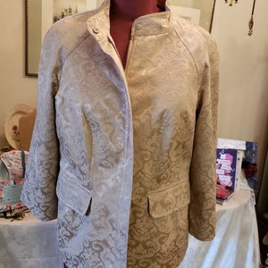May include: A gold-toned jacket featuring a tone-on-tone damask floral pattern, a high collar with a single button closure, a front zipper, and two flap pockets. The jacket is displayed on a dark red dress form, showcasing its structured fit.