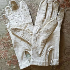 May include: A pair of off-white leather gloves with a textured surface. The gloves have a classic design with a slit at the wrist and are likely vintage. The gloves are displayed on a patterned surface.