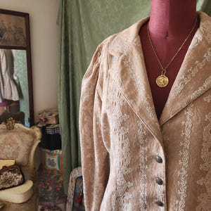 May include: A tan blazer featuring intricate white embroidered patterns, long sleeves with gathered shoulders, a notched lapel, and a front button closure with dark metallic buttons. A gold-toned pendant necklace rests on the neckline.