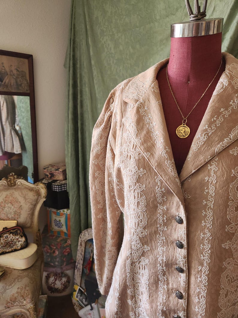 May include: A tan blazer featuring intricate white embroidered patterns, long sleeves with gathered shoulders, a notched lapel, and a front button closure with dark metallic buttons. A gold-toned pendant necklace rests on the neckline.