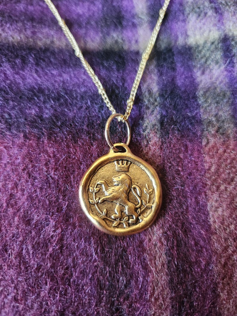 May include: A gold-toned circular pendant necklace featuring a raised relief of a lion standing on its hind legs beneath a small crown, framed by thin leafy branches, suspended from a delicate gold-toned metal chain link necklace.
